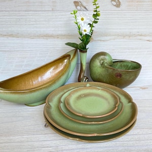 May include: A selection of green and brown ceramic tableware, including plates, bowls, and a vase with white flowers. The plates are stacked, and the bowls have unique shapes. The color palette is earthy and natural.