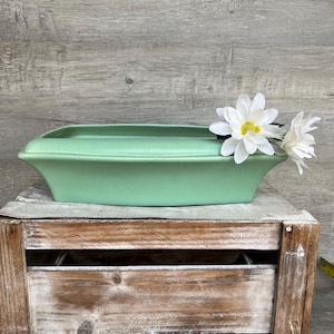 May include: A light green ceramic planter with a rectangular shape and rounded edges. The planter is sitting on a wooden crate and has white flowers on the edge.