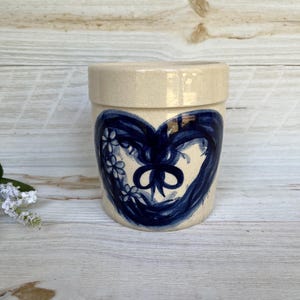 Könnte beinhalten: Ein beigefarbener Keramiktopf mit Deckel, verziert mit einem handgemalten blauen Herzdesign. Das Herz ist mit weißen Blumen und einer Schleife verziert. Der zylindrische Topf steht auf einer hellen Holzoberfläche.