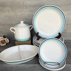 May include: A set of white ceramic dishes with blue and brown stripes. The set includes a large serving dish, a smaller serving dish, and several plates. The dishes are all in a rustic style.