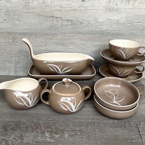 May include: A set of brown and white ceramic teacups, saucers, a creamer, a sugar bowl, and a gravy boat with a matching design. The set features a stylized leaf pattern in white on a brown background.