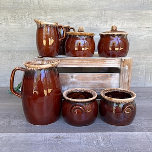 以下が含まれることがあります： 大きなピッチャー、小さなピッチャー、ふた付きのシュガーボウル2つ、取っ手付きの小さなボウル2つを含む、6つの茶色の釉薬をかけたセラミック製のキッチン用品のセット。すべてのアイテムは、茶色とクリーム色の釉薬で仕上げられています。