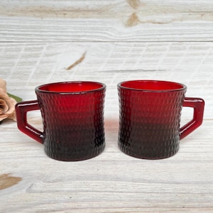 May include: Two ruby red glass mugs with textured surfaces and square handles. The mugs have a gradient effect, transitioning from a deep red at the base to a brighter red at the rim. The mugs are displayed on a light wood surface.