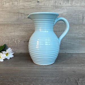 Puede incluir: Una jarra de cerámica azul claro con un diseño acanalado. La jarra tiene una boca ancha y un asa.
