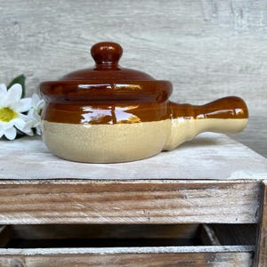 May include: A brown and beige ceramic pot with a lid and a handle. The pot is glazed and has a rustic look.