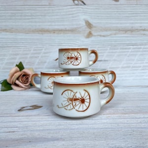 Pode incluir: Um conjunto de quatro canecas de cerâmica branca com borda e alça marrons. Cada caneca apresenta um desenho de roda de carroça marrom. As canecas estão dispostas sobre uma superfície clara de grão de madeira, com uma rosa e vegetação ao fundo.