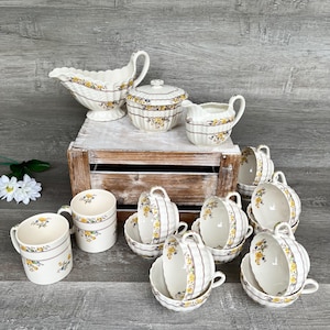 Puede incluir: Un conjunto de tazas de té, platillos, tazas, una salsera y una azucarera con tapa de porcelana blanca vintage. Cada pieza presenta un diseño floral amarillo con detalles en marrón y morado. El conjunto está dispuesto sobre una caja de madera.