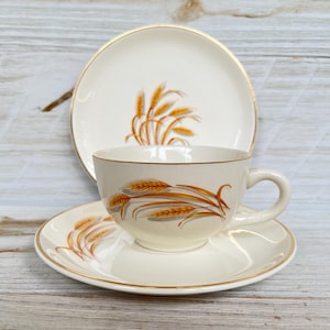 May include: A vintage cream-colored teacup, saucer, and plate set. The set features a wheat stalk design in shades of gold and brown. The edges of the cup, saucer, and plate are trimmed with a gold band. The set is displayed on a light wood surface.