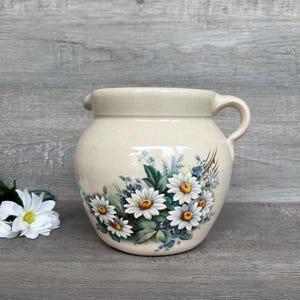 May include: A cream-colored ceramic pitcher with a floral design featuring white daisies and blue forget-me-nots. The pitcher has a rounded body and a handle.