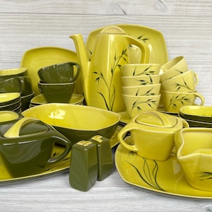 May include: A vintage ceramic tableware set in yellow and olive green. The collection features a teapot, cups, saucers, bowls, and serving dishes. Some pieces have black bamboo-style decoration. The arrangement is on a light wood surface.