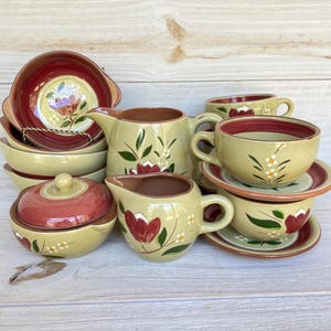 Puede incluir: Colección de vajilla de cerámica pintada a mano de estilo rústico. El juego incluye cuencos, tazas, platillos, una jarra de crema, un azucarero y una jarra. Las piezas son principalmente de color verde oliva con detalles burdeos y diseños florales.