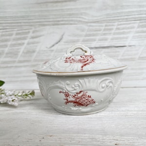 Puede incluir: Cuenco blanco de cerámica con tapa, con detalles en relieve ornamentados y un diseño floral rojo. El cuenco tiene un borde dorado y un asa en la tapa. El diseño general es vintage y decorativo.
