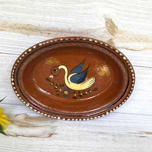 May include: Oval, hand-painted ceramic plate with a brown base and a decorative border. The plate features a central design of a swan with blue wings and yellow body, surrounded by blue and yellow floral accents.