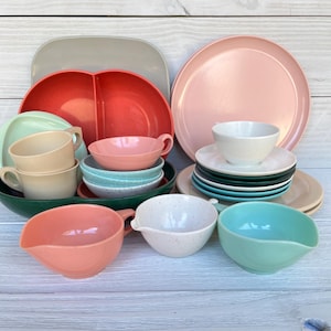 May include: A collection of colorful vintage dishware, including plates, bowls, and cups. The set features a variety of pastel colors such as pink, teal, and beige. The arrangement includes a divided serving dish and several saucers.
