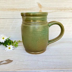 May include: A handcrafted ceramic mug with a green glaze. The mug has a cylindrical shape with a handle and a slightly flared rim. The color transitions from a darker green at the top to a lighter green and brown at the base. The mug is approximately 4 inches tall.