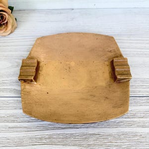 May include: A vintage, rectangular, bronze-colored tray with two small, rectangular handles on either side. The tray has a slightly curved edge and a textured surface, suggesting an aged appearance. The handles have a ridged design.