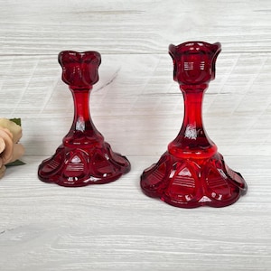 Pode incluir: Dois castiçais de vidro vermelho rubi. Cada um tem uma base detalhada e uma haste fina que leva a um topo em forma de taça. Os castiçais são feitos de vidro translúcido e brilhante.