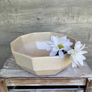 Peut inclure: Un bol octogonal en céramique beige avec une finition mouchetée. Le bol est rempli de fleurs blanches et repose sur une caisse en bois.