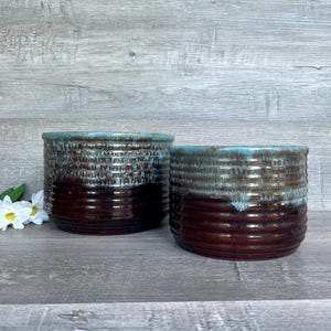 May include: Two brown and blue glazed ceramic pots with a textured, ribbed design. The pots are sitting on a light gray wood surface.