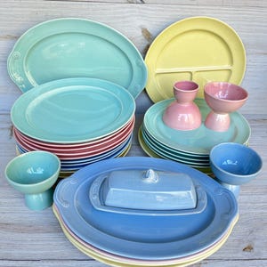 Puede incluir: Una colección de vajilla de cerámica colorida, que incluye platos, fuentes, cuencos y una mantequillera. Los platos son de varios colores como el turquesa, el amarillo, el rosa y el azul. La mantequillera tiene tapa y es de color azul claro. La vajilla está dispuesta sobre una superficie de madera clara.