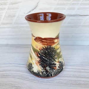 Puede incluir: Un jarrón de cerámica hecho a mano con forma acampanada. El jarrón presenta un diseño de paisaje con árboles negros y marrones sobre un fondo amarillo, naranja y verde. El borde y la base son marrones.