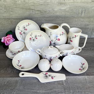 May include: A vintage set of white ceramic dishes with a pine cone and branch design in black and pink. The set includes a pitcher, creamer, sugar bowl, two salt and pepper shakers, a serving spoon, and four plates.