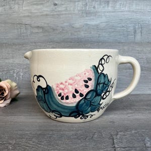 May include: A white ceramic jug with a hand-painted design of a watermelon slice in pink, green, and black. The jug has a handle and a spout.