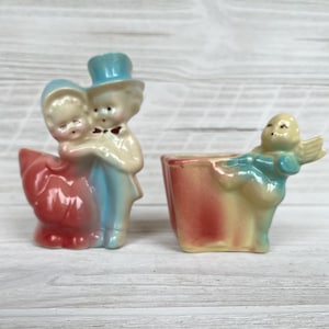 Könnte beinhalten: Zwei Vintage-Keramikfiguren. Eine zeigt ein Paar in formeller Kleidung, die andere einen Cherub. Die Figuren sind in Pastellfarben bemalt, darunter Rot, Blau und Gelb. Es sind wahrscheinlich Dekorationsartikel.