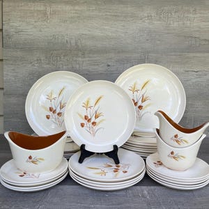 Könnte beinhalten: Ein cremefarbenes Vintage-Geschirrset mit einem Weizenähren-Design in Braun- und Goldtönen. Das Set umfasst Teller, Schalen und Saucieren. Die Teller sind gestapelt, und die Saucieren haben eine braune Innenseite.