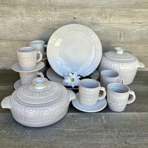 Peut inclure: Ensemble de vaisselle en céramique blanche, comprenant des assiettes, des tasses, des soucoupes et des plats de service. Les pièces présentent un motif texturé et en relief sur les bords et les couvercles. Une fleur blanche est présente.