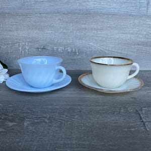 Pode incluir: Duas chávenas de chá vintage e pires. Uma é azul claro e a outra é branca com uma borda dourada.
