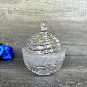 Peut inclure: Un pot en verre transparent avec un couvercle, orné d'un motif floral et d'un motif côtelé. Le pot est posé sur une surface en bois.