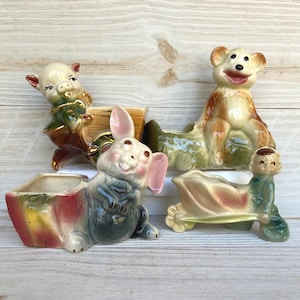 May include: Four vintage ceramic planters in the shape of animals. A pig in a cart, a bear, a bunny, and a boy pushing a flower cart. The planters are painted in pastel colors with detailed facial features and are ideal for small plants.