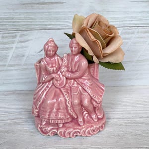Könnte beinhalten: Eine rosa Keramikvase mit einem sitzenden Paar in historischer Kleidung. Die Vase hat einen dekorativen Sockel und hält eine pfirsichfarbene Rose. Der Hintergrund ist eine helle Holzmaserung.