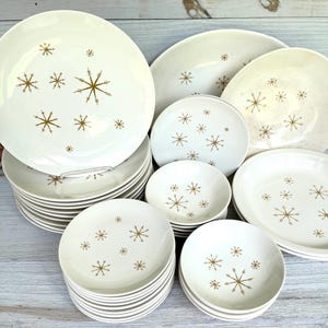 Peut inclure: Ensemble de vaisselle vintage en céramique blanche avec un motif d'éclats d'étoiles dorés. L'ensemble comprend des assiettes et des bols de différentes tailles, empilés et disposés sur une surface en bois clair. Les éclats d'étoiles dorés sont espacés.