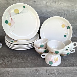 May include: White ceramic dinnerware set with a mid-century modern design. The set includes plates and cups, each decorated with a geometric pattern in yellow, green, and red. The plates are stacked, and the cups are arranged in a group.