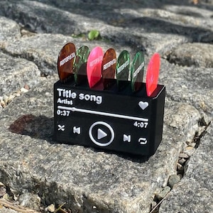 Może przedstawiać: Czarny uchwyt na kostki do gitary w kształcie odtwarzacza muzyki, z tekstem "Title song" i "Artist". Zawiera kilka kolorowych kostek do gitary. Kostki są w różnych kolorach, w tym czerwonym, zielonym i brązowym.