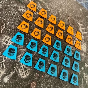 Può includere: Una vista dall'alto mostra una collezione di pedine. I pezzi sono a forma di scudo, con una parte superiore piatta e una inferiore a punta. Metà sono arancioni brillanti con un disegno a bersaglio nero, e l'altra metà azzurro chiaro con un disegno a teschio nero.