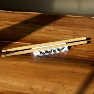 May include: Two wooden drumsticks resting on a white rectangular stand with the text "COLOGNE 07/10/11". The drumsticks are light brown with black tips. The stand is on a wooden surface.