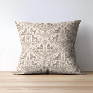 May include: Square decorative pillow with a beige and brown poodle pattern. The fabric features a repeating design of poodles in various poses, set against a damask-style background. The pillow is placed on a wooden surface.