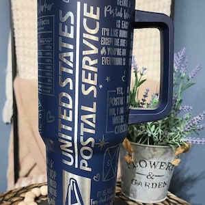 May include: A blue stainless steel tumbler with a silver lid and handle. It has a design of a mail truck, mailboxes, and the words "United States Postal Service" on the side. The tumbler also has the text "Being a Postal Worker is easy. It's like riding a bike except the bike is on fire. You're on fire. Everything is on fire. Yes, I am a Postal Worker. No, I don't know where the check is."