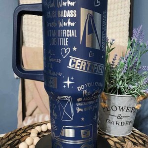 May include: A blue stainless steel travel mug with a black handle. The mug has a silver design with the text "Postal Worker", "Because Badass", "Miracle Worker", "Ain't an Official Job Title", "All the Games Until", "Misses a", "Certified", "Do You Know the Joke About the Unstamped Letter You Won't Get It", and a graphic of a mail truck.
