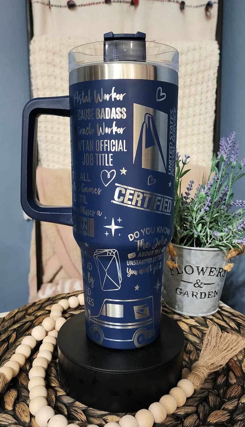May include: A blue stainless steel travel mug with a black handle. The mug has a silver design with the text "Postal Worker", "Because Badass", "Miracle Worker", "Ain't an Official Job Title", "All the Games Until", "Misses a", "Certified", "Do You Know the Joke About the Unstamped Letter You Won't Get It", and a graphic of a mail truck.