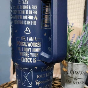 May include: A blue stainless steel tumbler with a handle. The tumbler has a silver design with text that reads "Being a Postal Worker is easy. It's like riding a bike except the bike is on fire. You're on fire. Everything is on fire. Yes, I am a Postal Worker. No, I don't know where your check is. Today's bad mood sponsored by blocked mailboxes. It is all fun and games until someone drops a tray of DPS."