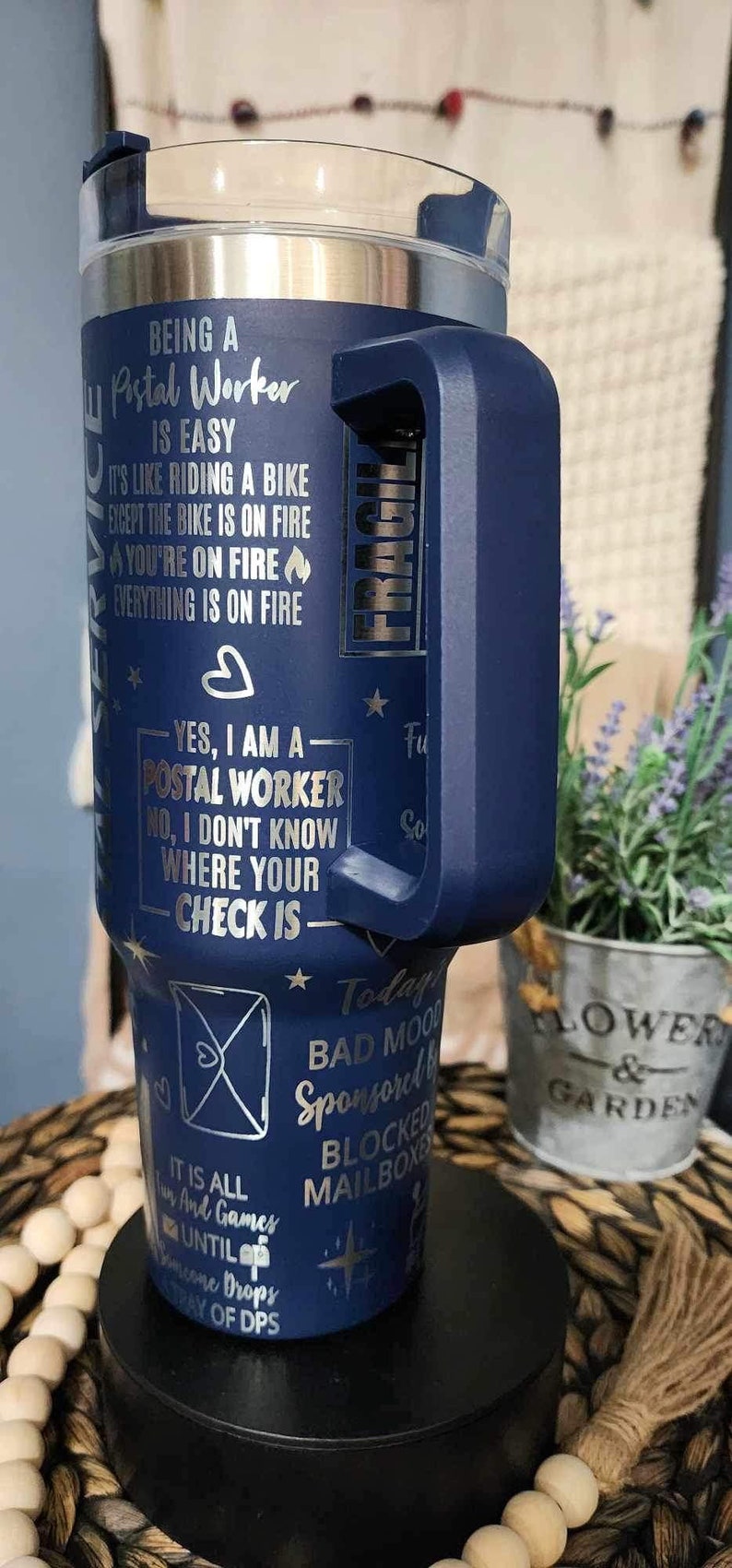 May include: A blue stainless steel tumbler with a handle. The tumbler has a silver design with text that reads "Being a Postal Worker is easy. It's like riding a bike except the bike is on fire. You're on fire. Everything is on fire. Yes, I am a Postal Worker. No, I don't know where your check is. Today's bad mood sponsored by blocked mailboxes. It is all fun and games until someone drops a tray of DPS."