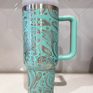 May include: A turquoise stainless steel travel mug with a silver paisley pattern and a handle. The mug has a lid.