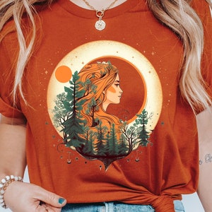 May include: A rust-colored t-shirt with a graphic of a woman's face surrounded by trees and a crescent moon. The moon is yellow with a red sun behind it.