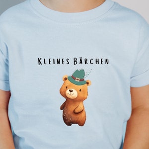 May include: A light blue t-shirt with a cartoon bear wearing a green hat. The text "KLEINES BÄRCHEN" is printed on the shirt.