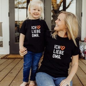 May include: A black t-shirt with the text "Ich Liebe Oma" and a pretzel heart graphic. A child wearing a matching t-shirt stands next to the adult.