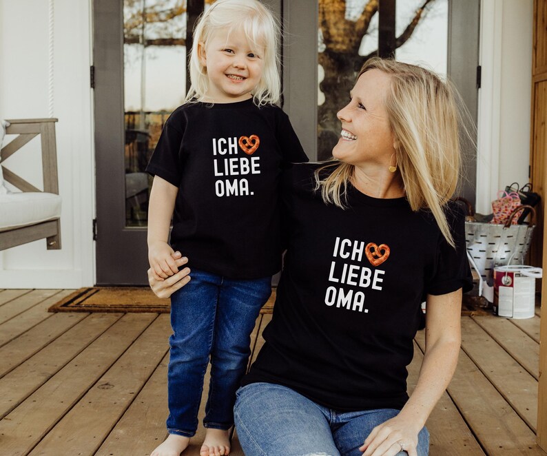 May include: A black t-shirt with the text "Ich Liebe Oma" and a pretzel heart graphic. A child wearing a matching t-shirt stands next to the adult.